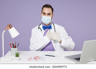 Male Cardiologist Doctor Man In White Medical Suit Face Sterile Mask Latex Gloves From Coronavirus Covid-19 Sit At Clinic Desk Put Folded Hand On Heart Isolated On Violet Background Studio Portrait