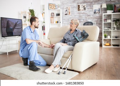 Male Care Taker In A Nursing Home Holding Arm Of Old Woman Sitting On The Couch. Big Tv In The Background.