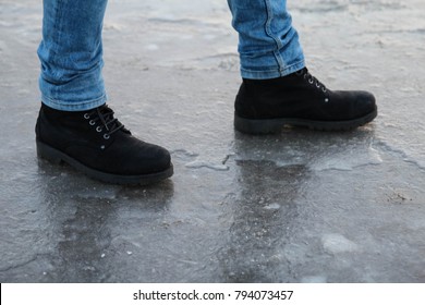 A Male Carefully Walking On The Slippery Road - Frozen Puddle Covered With Ice, Close Up View Of Legs In Black Shoes And Jeans. Danger Concept.