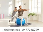 Male caregiver or doctor physiotherapist helping young man doing sport exercises sitting on fit ball in rehabilitation center helping patient in rehab. Physiotherapy and rehabilitation concept.