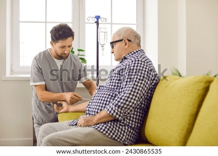 Male caregiver preparing elderly patient for IV therapy at home. Senior man sitting on sofa receiving intravenous treatment or vitamin therapy at home. Elderly people health care and medical support