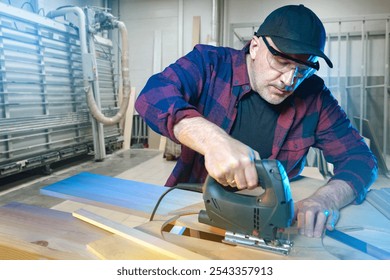 Male carpenter at work. Furniture maker uses electric jigsaw. Carpenter guy is focused on his work. Furniture maker cuts boards in factory workshop. Experienced male carpenter in checkered shirt - Powered by Shutterstock