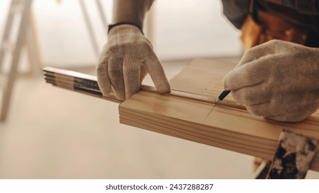 Male carpenter in work gloves measures and marks a wooden plank inside a home. This skilled worker brings expertise to upgrade the house's interior. He focuses on trim work, showcasing craftsmanship. - Powered by Shutterstock