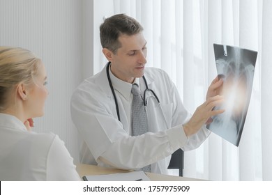 Male Caucasian Doctor Showing X-ray Film And Diagnose The Way To Treatment With Young Female Patient At Hospital Office, Light Effect, Selective Focus.