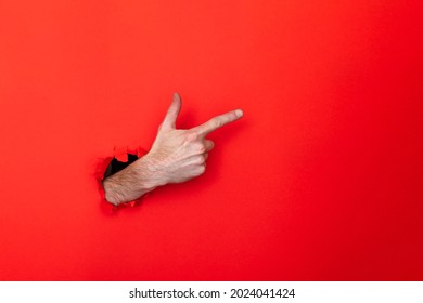 Male caucasian hand through a hole pointing against a red background