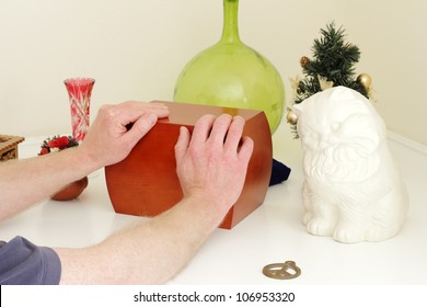 Male Caucasian Hands Holding A Wood Urn That Is On A White Mantle Surrounded By The Departed Loved Ones Mementos.