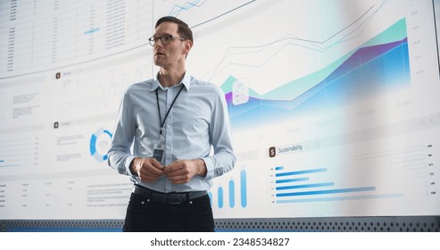 Male CEO Of Data Science Company Is Giving Presentation In Front Of Big Digital Screen With Graphs And Charts. Successful Caucasian Man Reviewing Business Accomplishments And Objectives In the Office.