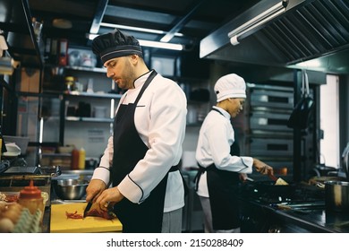 Male Chef Chopping Food While Cooking A Meal In The Kitchen At Restaurant. His Female Coworker Is In The Background.