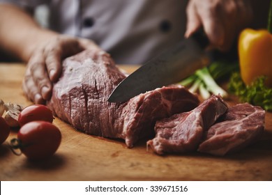 Male Chef Cutting Big Piece Of Beef On Wooden Board In Restaurant Kitchen. Man Cook Preparing Steak And Vegetable Garnish.    