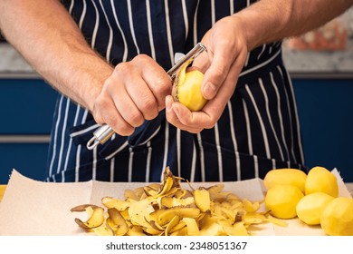 male chef hands peeling a bunch of potatoes with a food peeler in front of a striped cooking apron in the kitchen. preparing fresh vegetables on a wooden cutting board for cooking a healthy meal. - Powered by Shutterstock