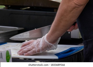 A Male Chef Prepares Food In A Kitchen Restaurant. He Has Clear Plastic Gloves On His Hands And A Large Stainless Steel Knife Cutting Onion On A White Plastic Board.