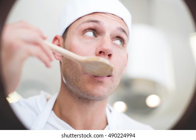 Male Chef Tasting Food From Saucepan In Commercial Kitchen