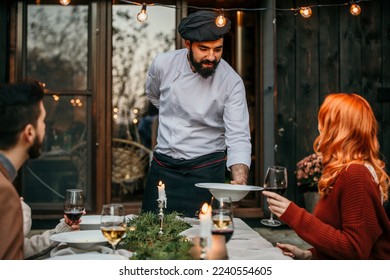 A male chef in uniform is serving a private elegant dinner to a group of people, Focus on a chef putting down the plate to a redhead woman - Powered by Shutterstock