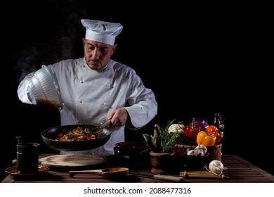 Male chef in white uniform pouring boiled spaghetti into pan wok for cooking pasta with vegetables. Backstage of preparing traditional italian dish on black background