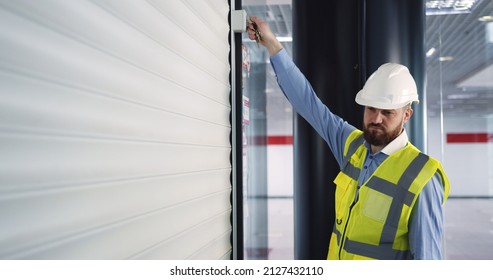 Male Chief Engineer In Hardhat And Vest Standing Inside Commercial Building Construction Site, Closing An Automatic Roller Door Before Leaving Work.