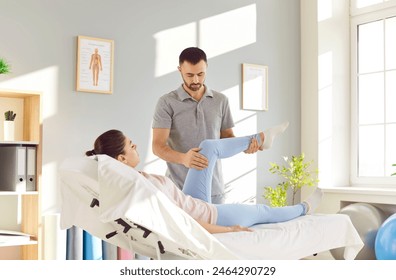 Male chiropractor or physiotherapist working with female patient. Man doctor standing by medical bed and examining leg of young woman who complains about knee pain and asks for physiotherapy treatment - Powered by Shutterstock