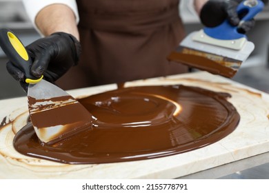 Male chocolatier uses a spatula to stir the tempered liquid chocolate on a granite table - Shutterstock ID 2155778791