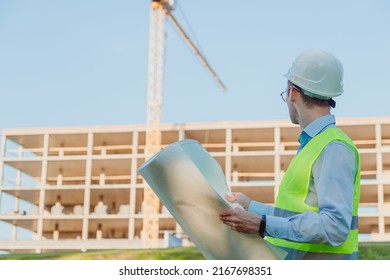 Male Civil Engineer In Helmet And Vest, Checking Architectural Drawing At Construction Site. Industrial Supervision In Construction