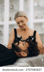 Male client lying down while cosmetologist applies permanent makeup to his eyebrows, enhancing facial features at beauty salon