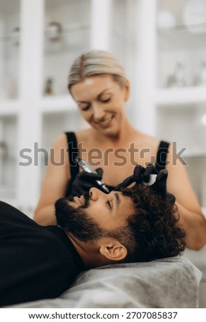 Male client lying down while cosmetologist applies permanent makeup to his eyebrows, enhancing facial features at beauty salon