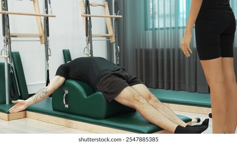 Male client practicing leg lifts on Pilates arc barrel with instructor assistance for improving core stability, leg strength, and flexibility at gym. Student lying and posing teaser pose. Habituate. - Powered by Shutterstock