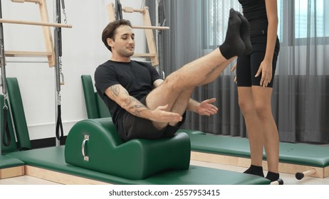 Male client practicing leg lifts on Pilates arc barrel with instructor assistance for improving core stability, leg strength, and flexibility at gym. Student lying and posing teaser pose. Habituate. - Powered by Shutterstock