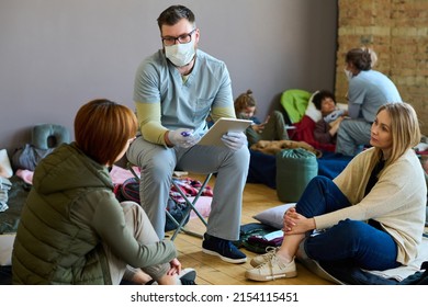 Male Clinician In Uniform And Mask Giving Medical Advice To One Of Young Female Refugees Sitting On Sleeping Place On The Floor Of Camp