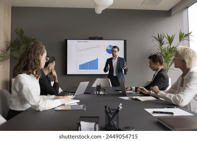 Male coach hold seminar for staff, makes presentation, provides sales statistics explanation gathered in modern conference room, with monitor screen with infographic charts, graphs and diagrams data