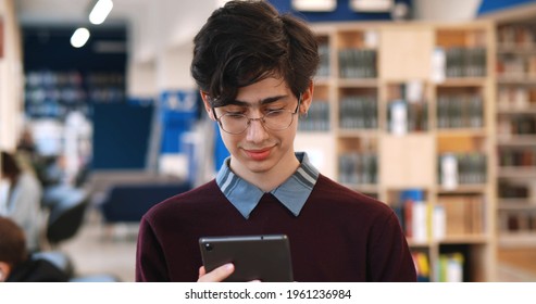 Male College Student Using Digital Tablet Standing In Library. Portrait Of Young Man In Eyeglasses Reading Ebook In Tablet Pc Studying In University Library