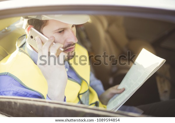 Male construction engineer in white helmet, shirt\
and yellow waistcoat sits in the car and scans documents while\
talking on the phone