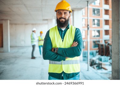 A male construction worker with a beard stands confidently at a construction site, arms crossed, wearing a hard hat and high-visibility vest, as colleagues work in the background. - Powered by Shutterstock