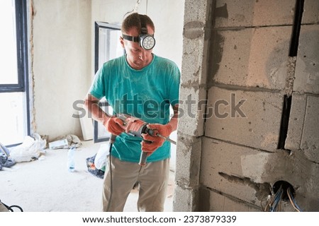 Male construction worker drilling brick wall with electric drill in apartment under renovation. Man wearing headlamp flashlight and work gloves while using professional drilling tool at home.