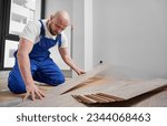 Male construction worker laying laminate wood plank on floor underlayment in living room. Bearded man in work overalls installing laminate timber flooring in apartment under renovation.