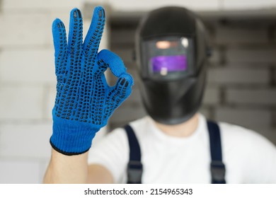 Male construction worker wearing a welding helmet and blue gloves shows the ok sign.