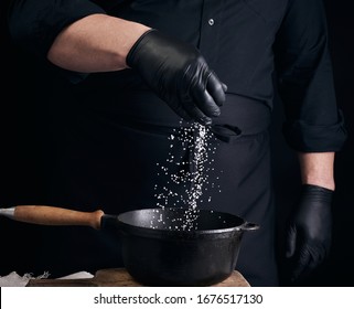 Male Cook In Black Uniform And Latex Gloves Salt Food In A Black Cast-iron Pan, Salt Crystals Froze In The Air, Low Key