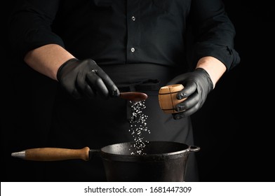 Male Cook In Black Uniform And Latex Gloves Salt Food In A Black Cast-iron Pan, Salt Crystals Froze In The Air, Low Key