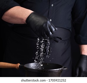 Male Cook In Black Uniform And Latex Gloves Salt Food In A Black Cast-iron Pan, Salt Crystals Froze In The Air, Low Key