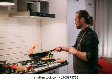 A Male Cook Is Cooking At The Stove At Home In The Kitchen. A Young Guy In An Apron In The Kitchen At The Gas Stove Fries Food In A Wok