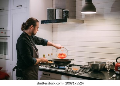 A Male Cook Is Cooking At The Stove At Home In The Kitchen. A Young Guy In An Apron In The Kitchen At The Gas Stove Fries Food In A Wok