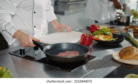 Male Cook Preparing Pork Meat In Frying Pan With Olive Oil On Stove, Cooking Delicious Meal. Man Working In Professional Kitchen Cooking Roast Beef Steak For Authentic Dish Recipe. Close Up.