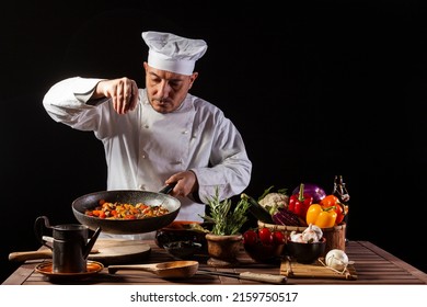 Male cook in white uniform and hat putting salt and herbs on food plate with vegetables before serving while working in a restaurant kitchen