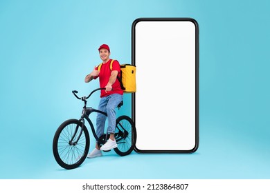 Male Courier On Bike Near Big Empty Phone Screen Gesturing Thumbs Up Approving Food Delivery App On Blue Studio Background, Wearing Uniform And Cap, Carrying Yellow Backpack Bag. Mockup