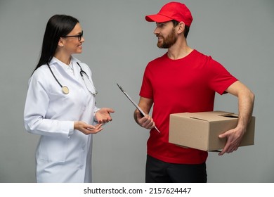 Male Courier In Red Uniform Handing Cardboard Parcel To Female Client Doctor, Carrying Parcel With Goods Ordered Online. Indoor Studio Shot Isolated On Grey Background 