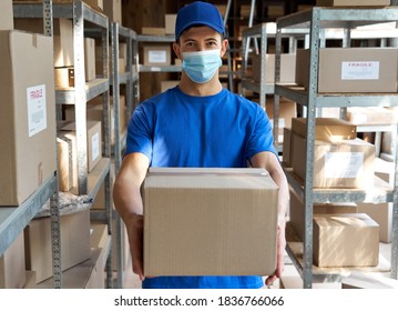 Male courier worker wearing uniform and face mask holding parcel box giving package to camera standing in stock warehouse, portrait. Fast express safe delivery during covid 19 pandemic concept. - Powered by Shutterstock