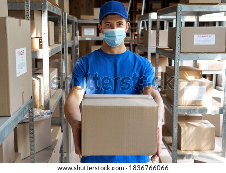 Male courier worker wearing uniform and face mask holding parcel box giving package to camera standing in stock warehouse, portrait. Fast express safe delivery during covid 19 pandemic concept.