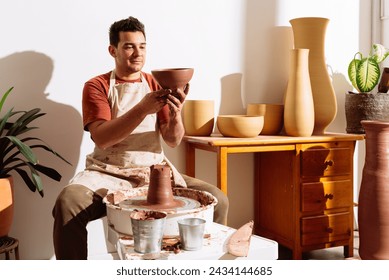 Male craftsman in apron sitting near table with vases and bowls while working on clay vase in pottery studio - Powered by Shutterstock