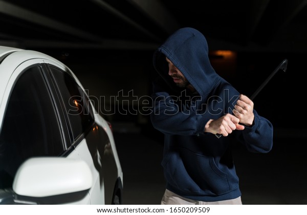Male criminal breaking car window with crowbar in\
dark parking lot