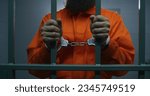 Male criminal in orange uniform and handcuffs holds metal bars, stands in prison cell, looks at camera. Prisoner serves imprisonment term in jail. Detention center or correctional facility. Portrait.