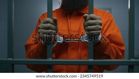Male criminal in orange uniform and handcuffs holds metal bars, stands in prison cell, looks at camera. Prisoner serves imprisonment term in jail. Detention center or correctional facility. Portrait.
