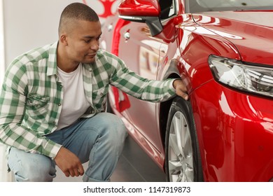 Male Customer Examining Tires Of A New Automobile On Sale. Handsome African Man Choosing New Car At The Dealership Salon. Attractive Male Driver Buying An Auto. Quality, Safety, Technology Concept